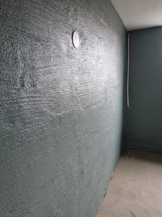 Rumshörn med texturerade gråa väggar och en liten vit ventilationsskiva.