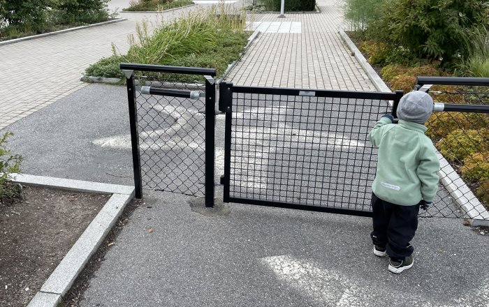 Barn i mössa står vid öppet svart stängsel i en lekpark.