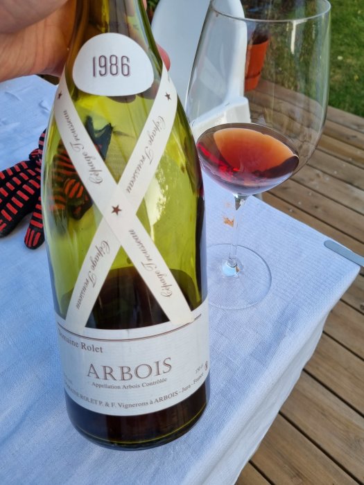 Flaska vinetikett "Domaine Rolet Arbois 1986" och ett glas med rödvinsfärgad dryck på ett bord med vit duk.