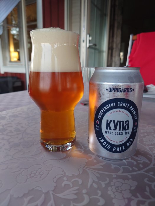 Ett glas fyllt med Oppigårds West Coast IPA bredvid en burk av samma öl på ett broderat bord.