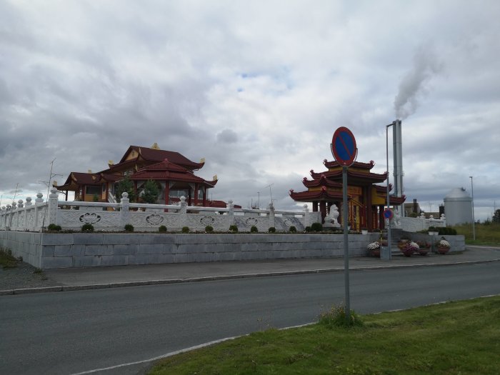 Traditionellt asiatiskt tempel med röda tak och vita väggar vid en väg, mot molnig himmel i Trondheim.