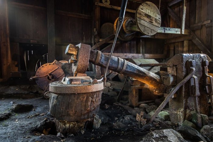 Gammal smedja med anvil på stock, rörliga delar och sparkad jordgolv belyst av solljus.