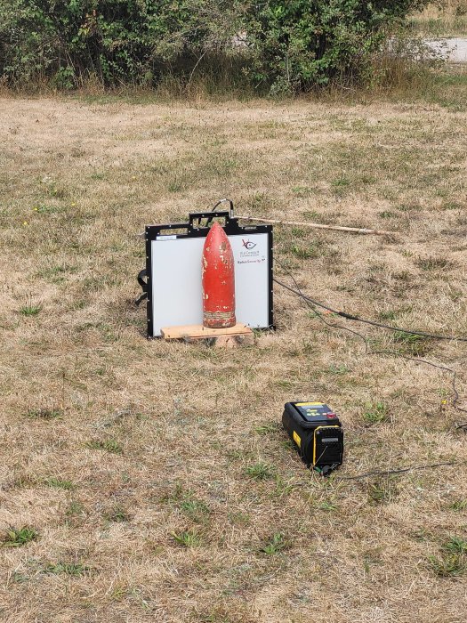 En kartong med röd snubbeltråd placerad på en äng med en bombdetektor bredvid.