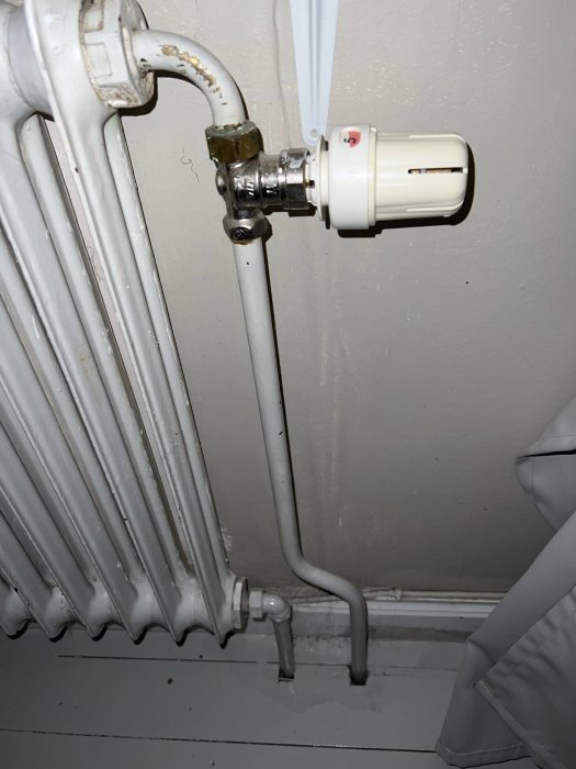 Vit elementradiator med termostatventil och anslutna rör mot en grå vägg.