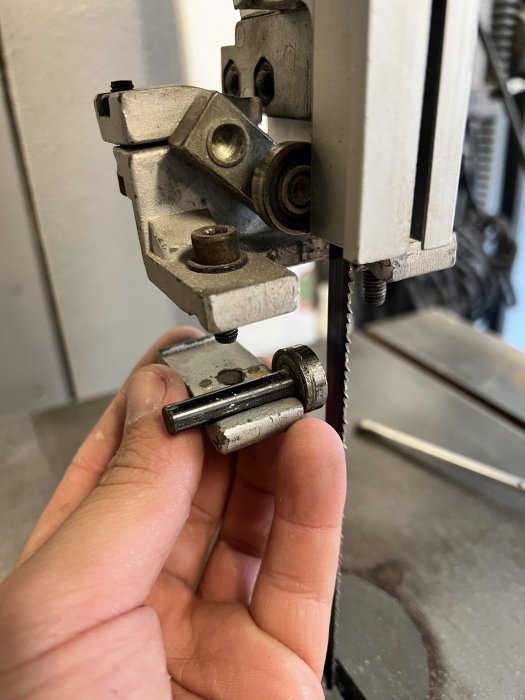 Hand håller ett sprucket metallhus som är en del av en större mekanism, nära en bandsåg.