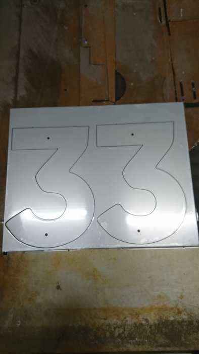 Metallplåt med utskurna delar formade som siffran 33 placerade på ett trägolv.