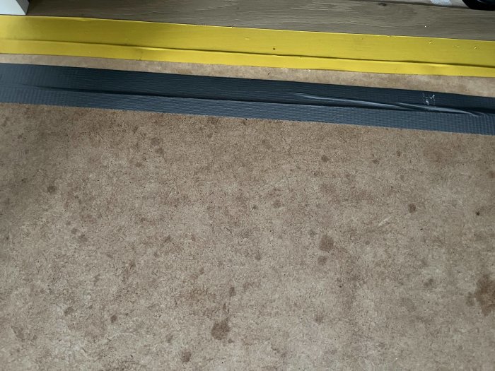 Gul skyddstejp på nytt golv vid en tröskel, tejpens kant inte helt fäst vid underlaget.