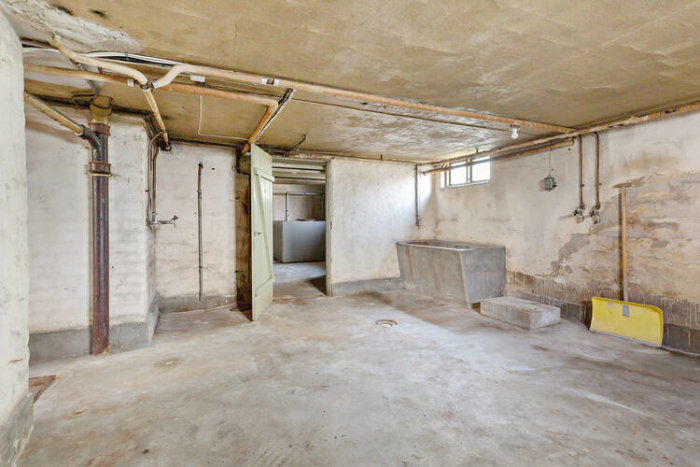 Oinredd källare med synliga rör och betonggolv som potentiellt renoveringsprojekt.