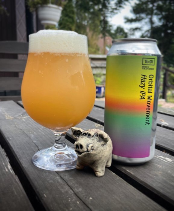 Ölglas fyllt med grumlig öl och en färgglad burk märkt "Hazy IPA", intill en keramikgris, på ett träbord.