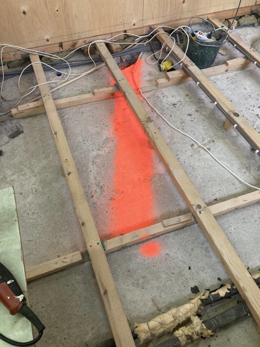 Avbilat golvområde markerat med neonorange för avloppsinstallation, omgivet av träreglar och elledningar.