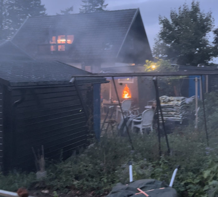 Dimmig vy av en trädgård med en brinnande eldstad, skjul och ett hus i bakgrunden på kvällen.