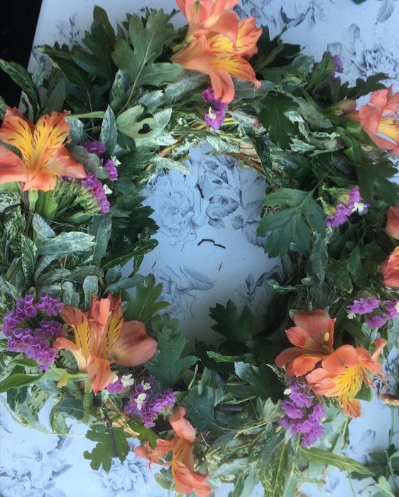 Handgjord krans med gröna blad och orangea blommor på en mönstrad bakgrund.