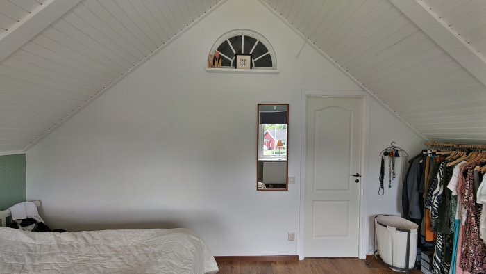 Renoverat sovrum i vitt med snedtak, dörr, fönster och klädstång.