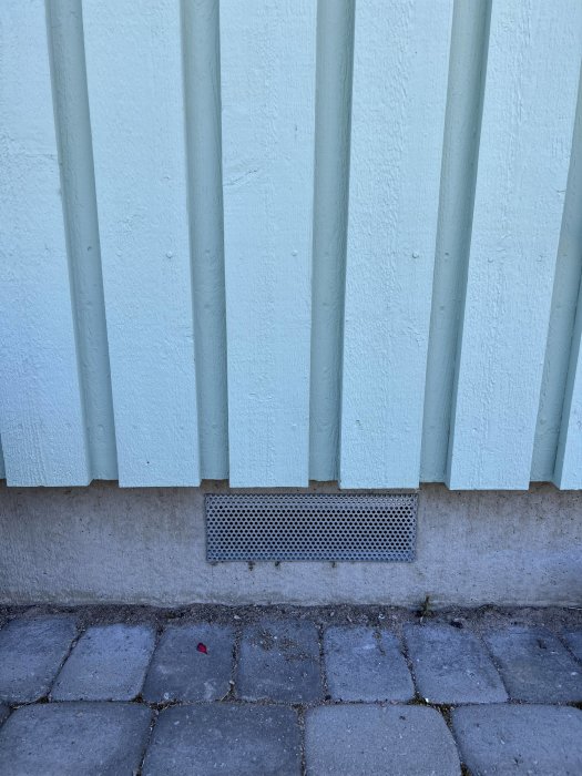 Stängd ventilationsgrill på en husgrund med isolerad krypgrund, intill en fasad och stenläggning.
