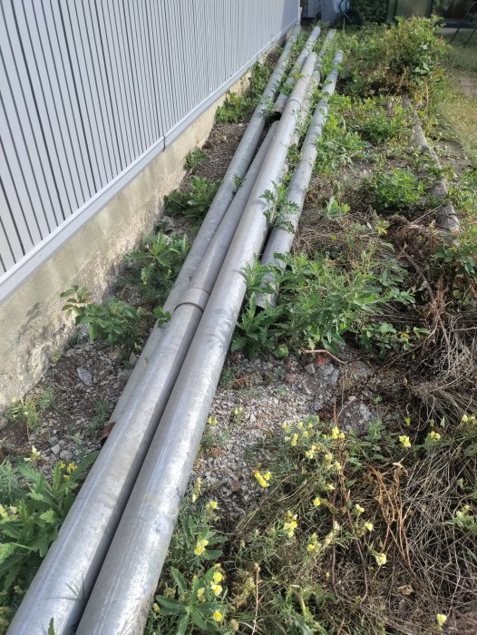 Rörledningar längs en byggnads vägg med omgivande vegetation.