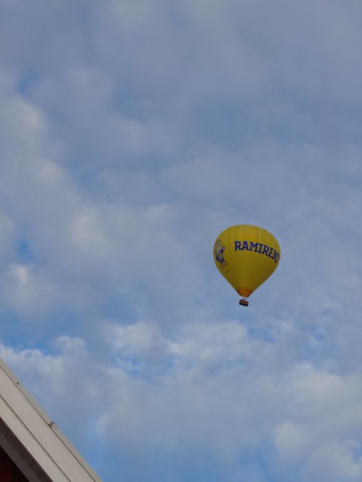 Gul varmluftsballong med texten "RAMIRENT" flyger över takkant mot molnig himmel.