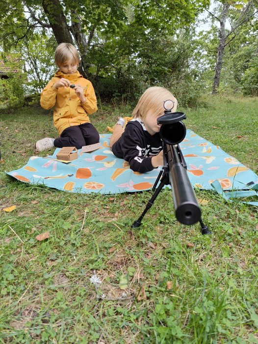 Två barn sitter på en filt i en lummig trädgård, det ena barnet siktar med en leksakspistol.