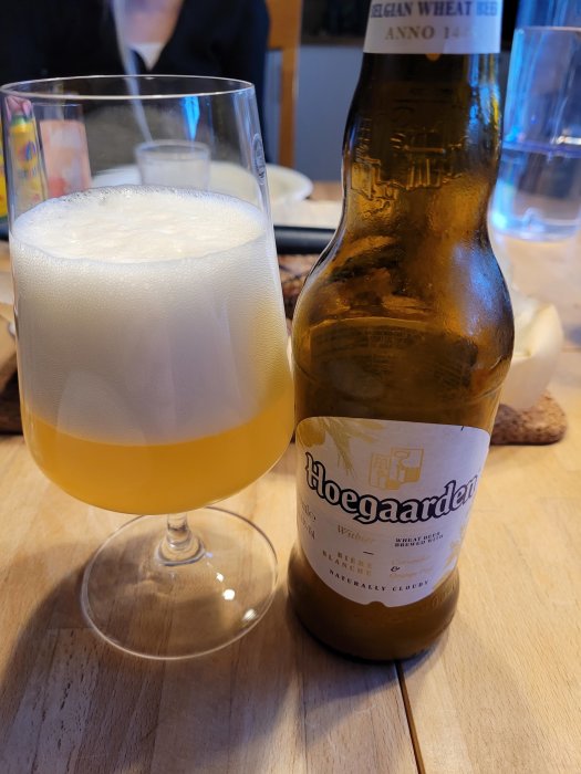 Veteöl i glas med skumkrona bredvid en öppnad Hoegaarden-flaska på bord.