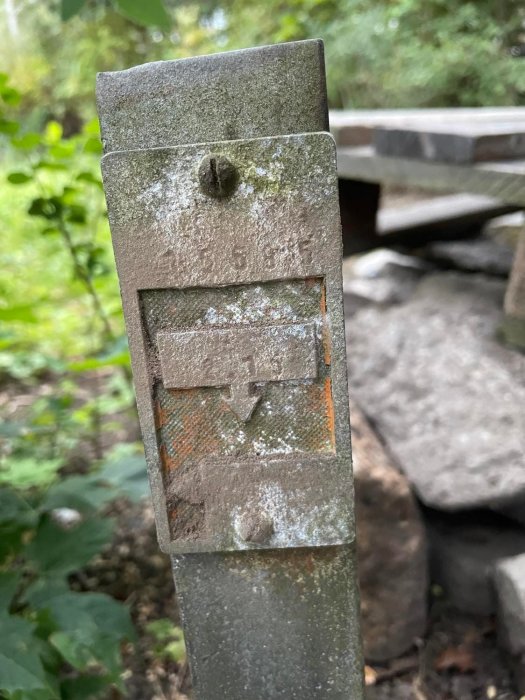 Närbild på en möglig metallstolpe med en skadad rustik ljushållare mot en suddig naturbakgrund.