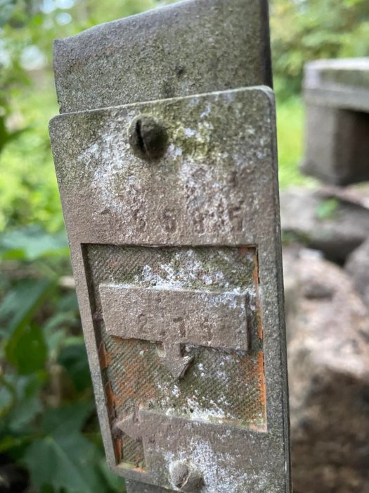 Närbild på en grå, mossa- och lavtäckt metallbeslag med skruvhål, monterad utomhus med suddig bakgrund.