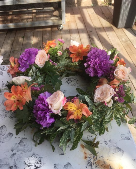 Handgjord blomsterkrans med rosor, alstroemeria och lila blommor på ett bord.