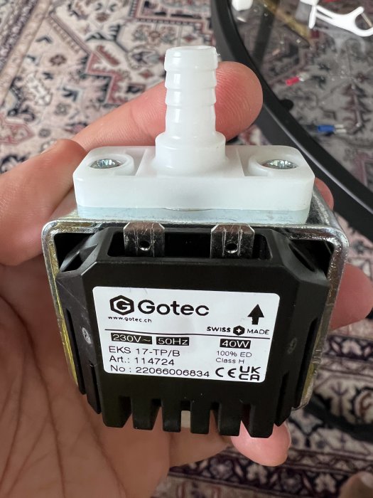 Hand håller en Gotec EKS 17-TP/B kondenspump mot en bakgrund av ett mönstrat tyg.