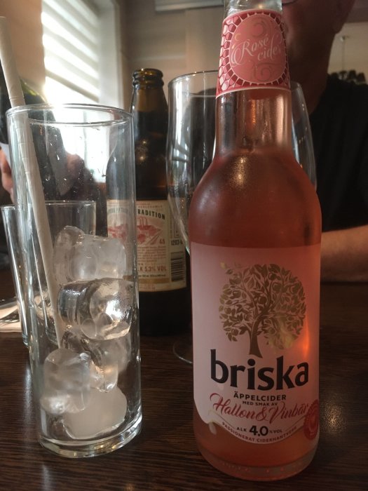 Flaska Briska rosécider med hallon- och vinbärssmak bredvid ett tomt glas med is, i bakgrunden syns del av en ölflaska.