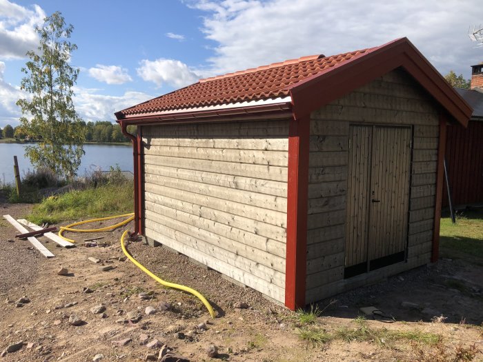 Renoverat fristående skjul med rött tak och nymonterade vindskivor, hängrännor och ljus panelvägg vid sjö.