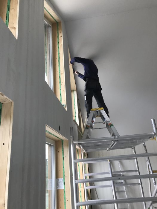 Person monterar panel runt fönster, står på stege med verktygsbälte inne i byggnad under konstruktion.
