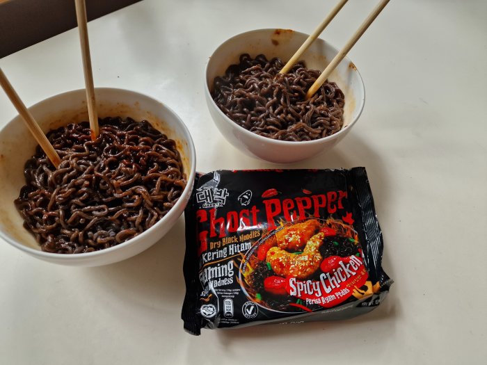 Två skålar med mörka nudlar och ätpinnar, tom Ghost Pepper-nudelpaket på ett ljust bord.