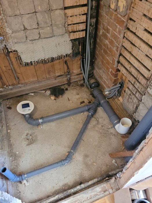 Rörinstallation i öppet golv med synliga bärverk och tegelvägg under renovering.