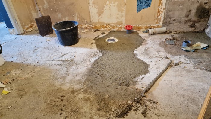 Nybetongat källargolv med lagat hål, hink och spade, och rörstumpar på golvet.