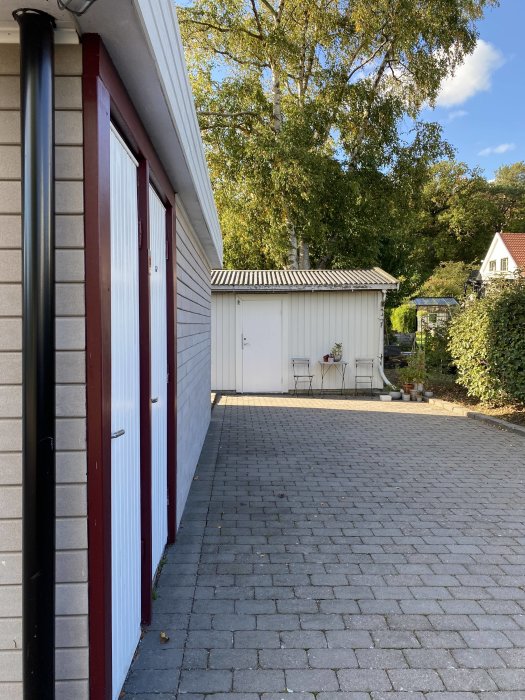 Uteplats med stenläggning och vit förrådsbyggnad vid en husvägg under en klar himmel.