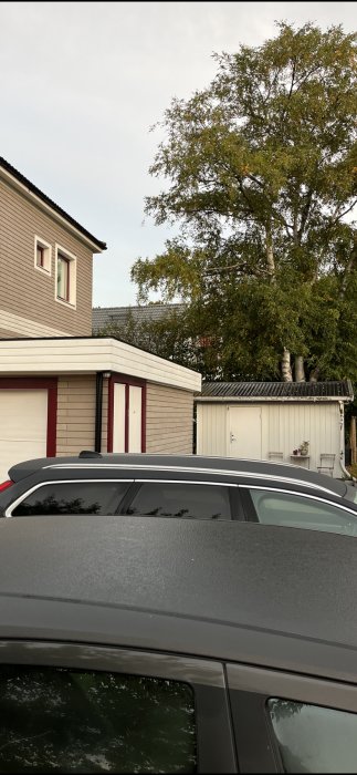 Bil och garage framför ett hus med träfasad och röda detaljer, omgivet av träd.