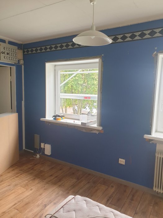 Renoverad rum med blåa väggar, trägolv och fönster med utsikt mot träd.
