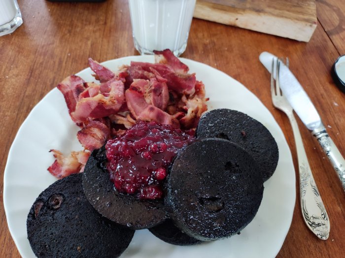 Tallrik med blodpudding toppat med lingonsylt och knaperstekt bacon, bredvid ett glas mjölk.