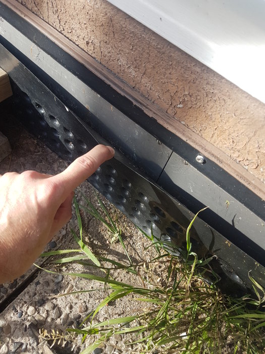 Hand pekar på fuktskydd vid husgrund med synliga fastsättningsdetaljer och en vägg i slamfärg.