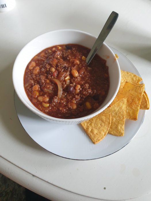 Hemlagad chili con carne i vit skål med sked och nachochips på vit tallrik.