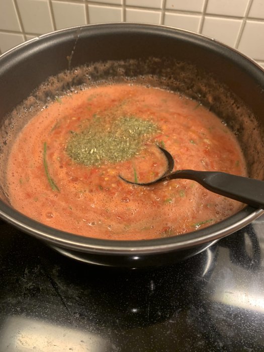 En gryta med kokande tomatsås och örter på ett spis, med en slev i såsen.