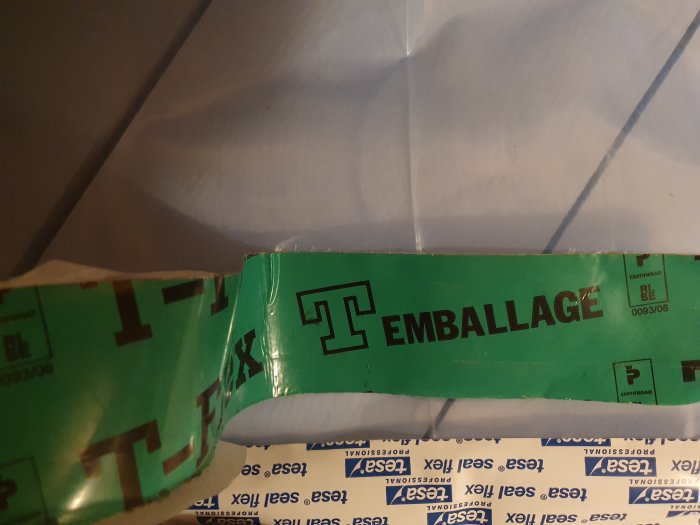 Grön tejp märkt med "TEMBALLAGE" fäst på en yta med pappersskikt under.