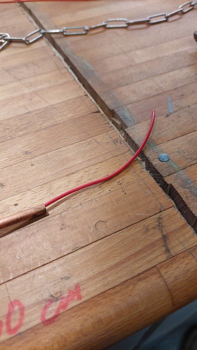 Röd elkabel som går genom ett kopplingselement på ett trägolv med synliga måttmarkeringar.
