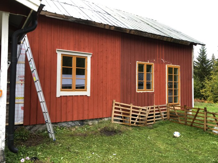 Röd stuga med nyinstallerade fönsterfoder och en stege lutad mot fasaden.