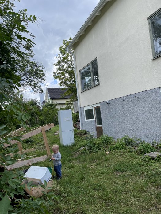 Ett barn står i en trädgård bredvid en öppen kyl som utställs, med nybyggt trästaket och husfasad i bakgrunden.