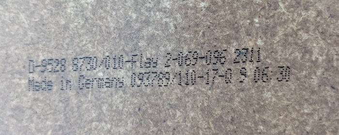 Närbild av en suddig etikett med punktmatrisutskrift som visar text och siffror på en pappartad yta.