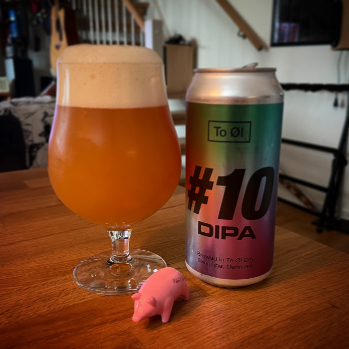 Ölglas fyllt med en dimmig dryck bredvid en burk märkt "#10 DIPA" och en liten rosa plastgris på ett bord.