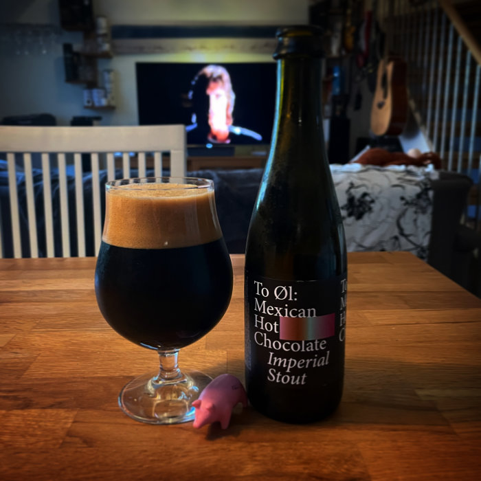 Mörkt öl i glas bredvid flaska märkt "To Øl Mexican Hot Chocolate Imperial Stout", i bakgrunden en tv och gitarr.