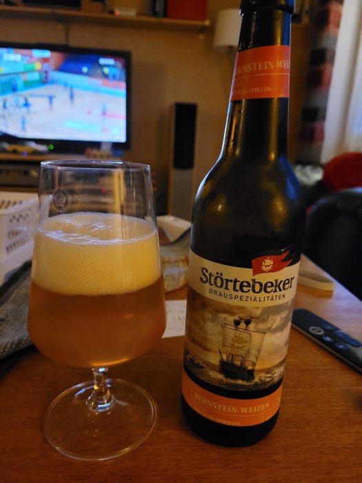 Flaska och glas med veteöl framför en TV som visar sport.