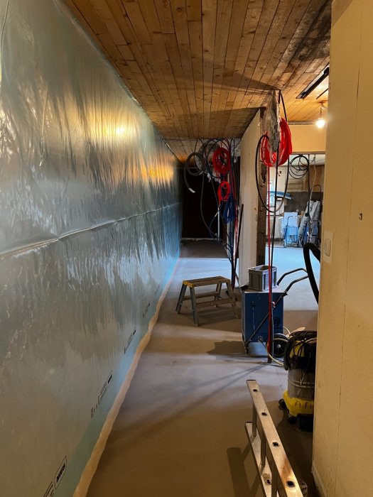 Arbetsrum med upphängd plast i taket, byggmaterial på golvet och verktyg längs väggen.