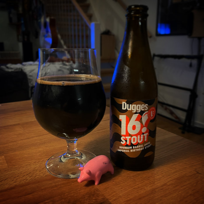 Ölglas fyllt med stout bredvid en flaska Dugges 16% Stout och en liten rosa plastgris på ett bord.