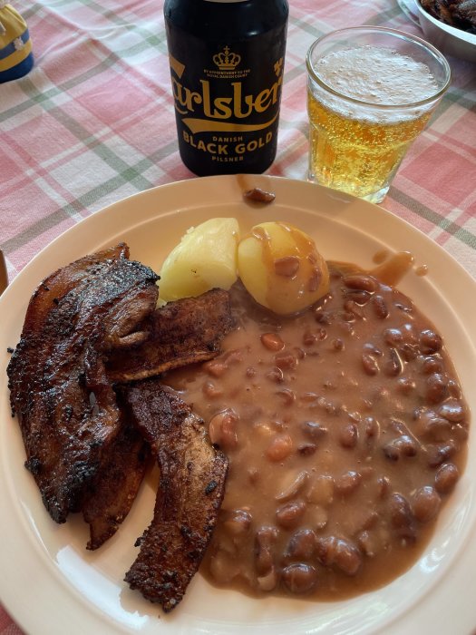 En tallrik med stekt fläsk, kokta potatisar och bruna bönor, bredvid ett glas öl och en flaska Carlsberg.